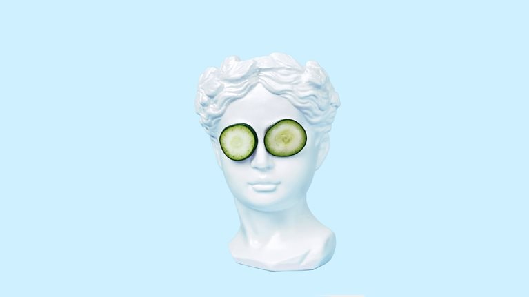 A bust of a young woman, inspired by ancient Greek art, wearing cucumber slices on her eyes and set against a light blue backdrop.