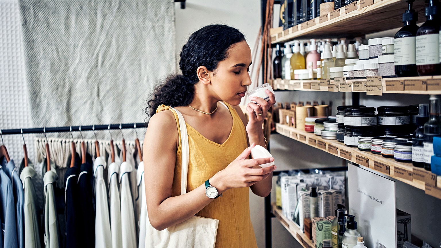 A woman is smelling a skin care product while standing in front of a fully stocked shelf at an organic store.