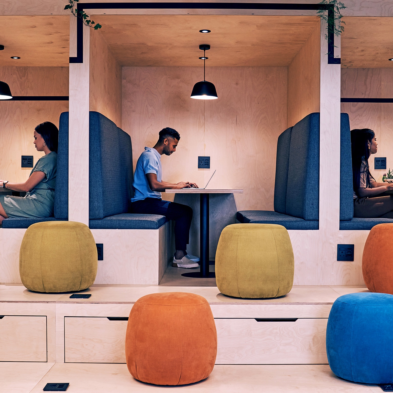 Three individuals seated in separate modern office booths, each focused on their laptops. The minimalist design features light wood, blue upholstery, and colorful ottomans, creating a functional and stylish workspace.