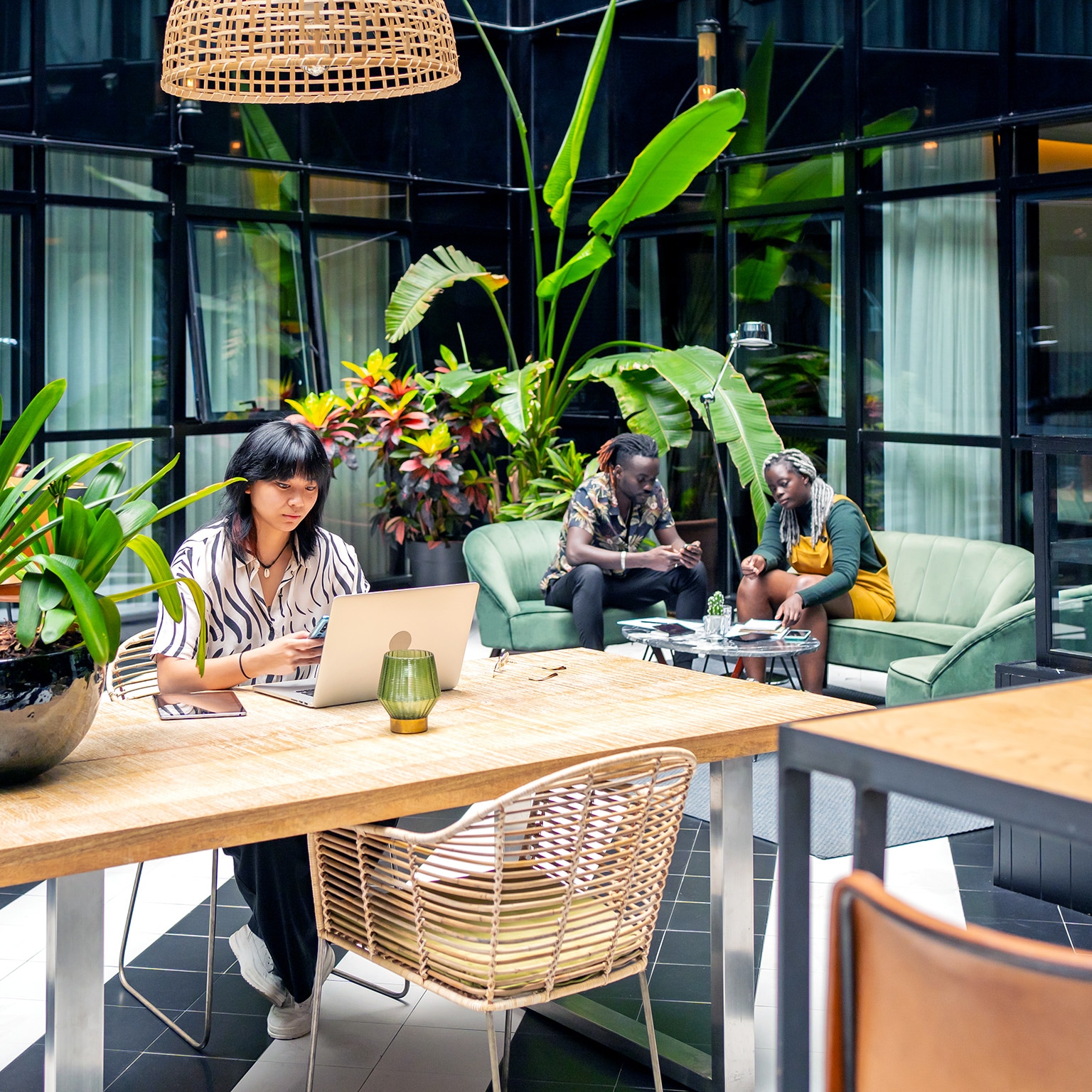 In the foreground, a woman is busy on her laptop, while two individuals seated on green armchairs are engaging in collaboration in the background. The well-lit shared space boasts expansive windows and is adorned with numerous sizable plants.