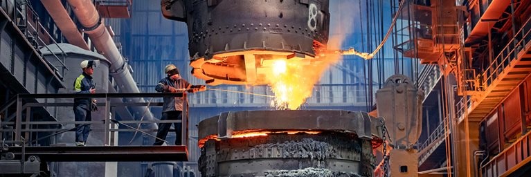 Steelworkers in heat protective gear starting a glowing molten steel pour in a steelworks factory.